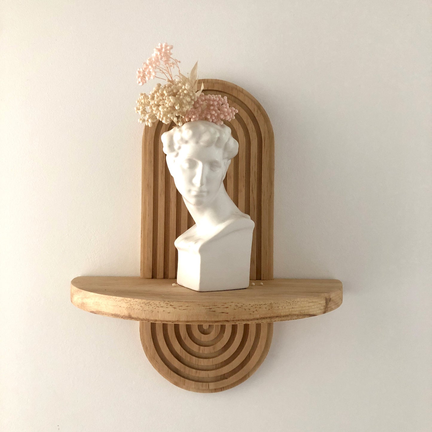 Wooden wall-mounted shelf with an arched design, holding a white bust sculpture adorned with dried flowers. The shelf's natural wood finish and minimalist design make it perfect for modern home decor.
