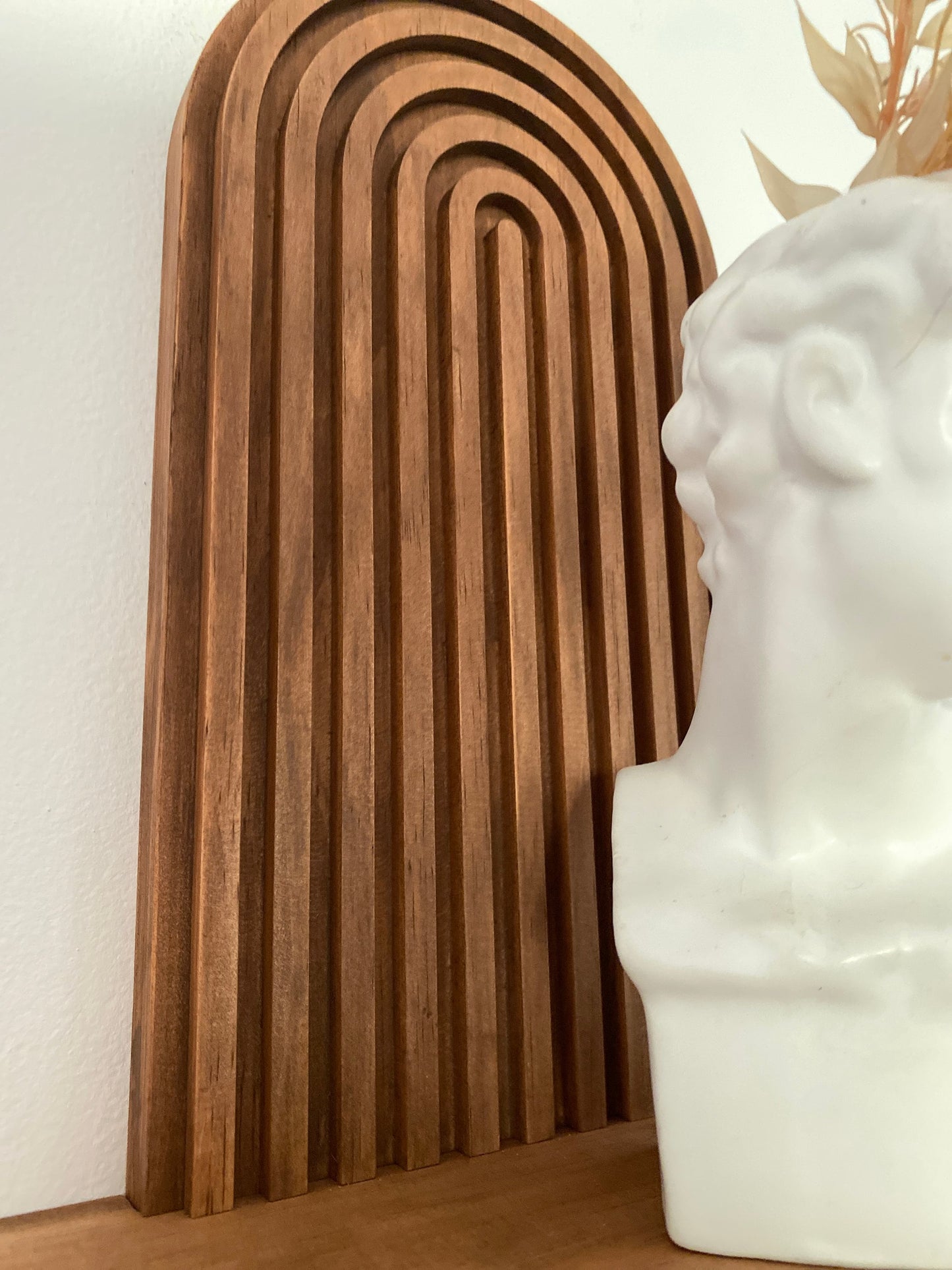 Close-up of a wooden wall-mounted shelf with an arched design, featuring a white bust sculpture adorned with dried flowers. The image highlights the intricate craftsmanship and natural wood finish of the shelf.
