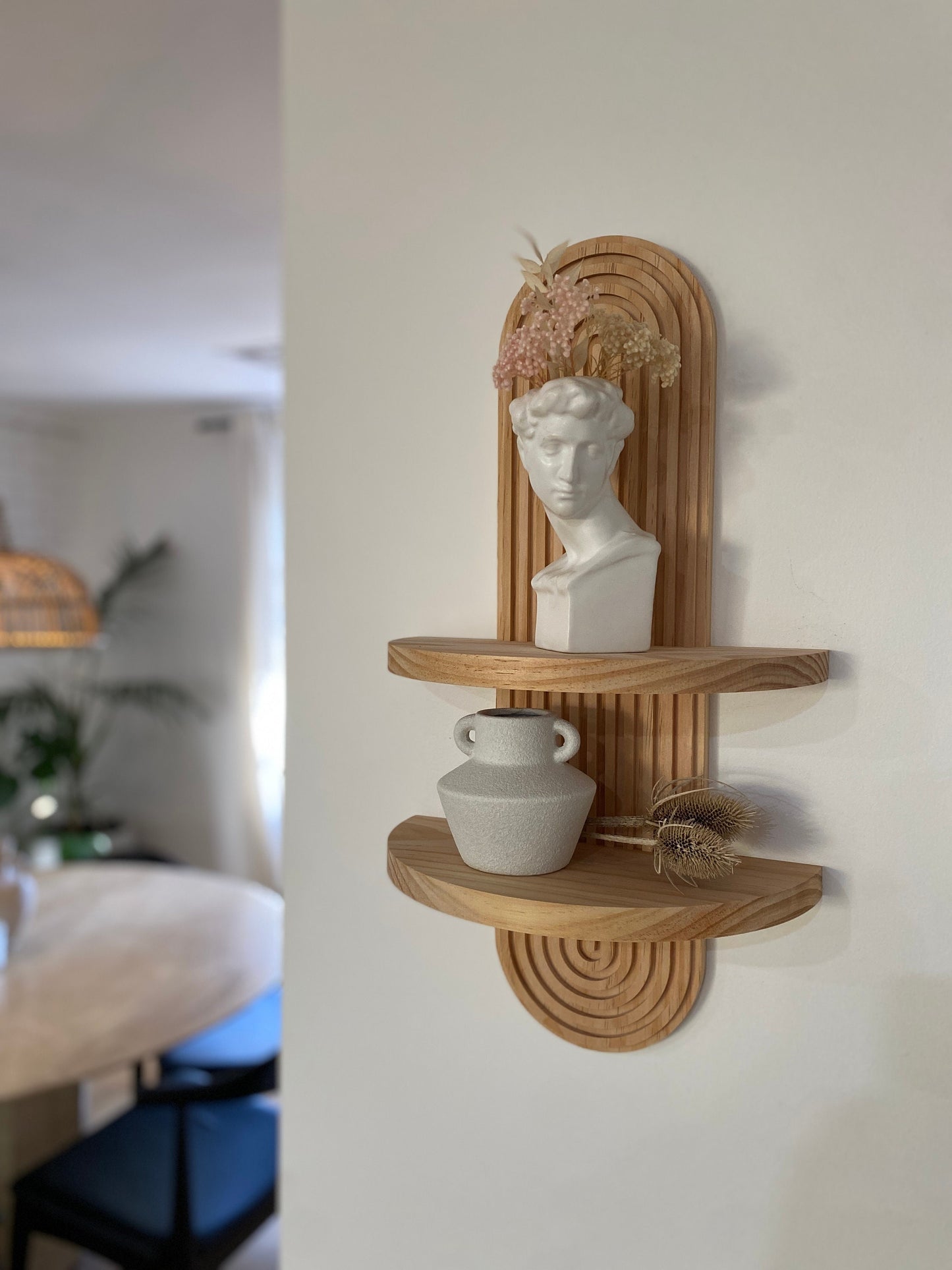 Wooden wall-mounted shelf with an arched design featuring two tiers. The top shelf holds a white bust sculpture with dried flowers, and the bottom shelf holds a ceramic vase and decorative items. Displayed in a modern living space.