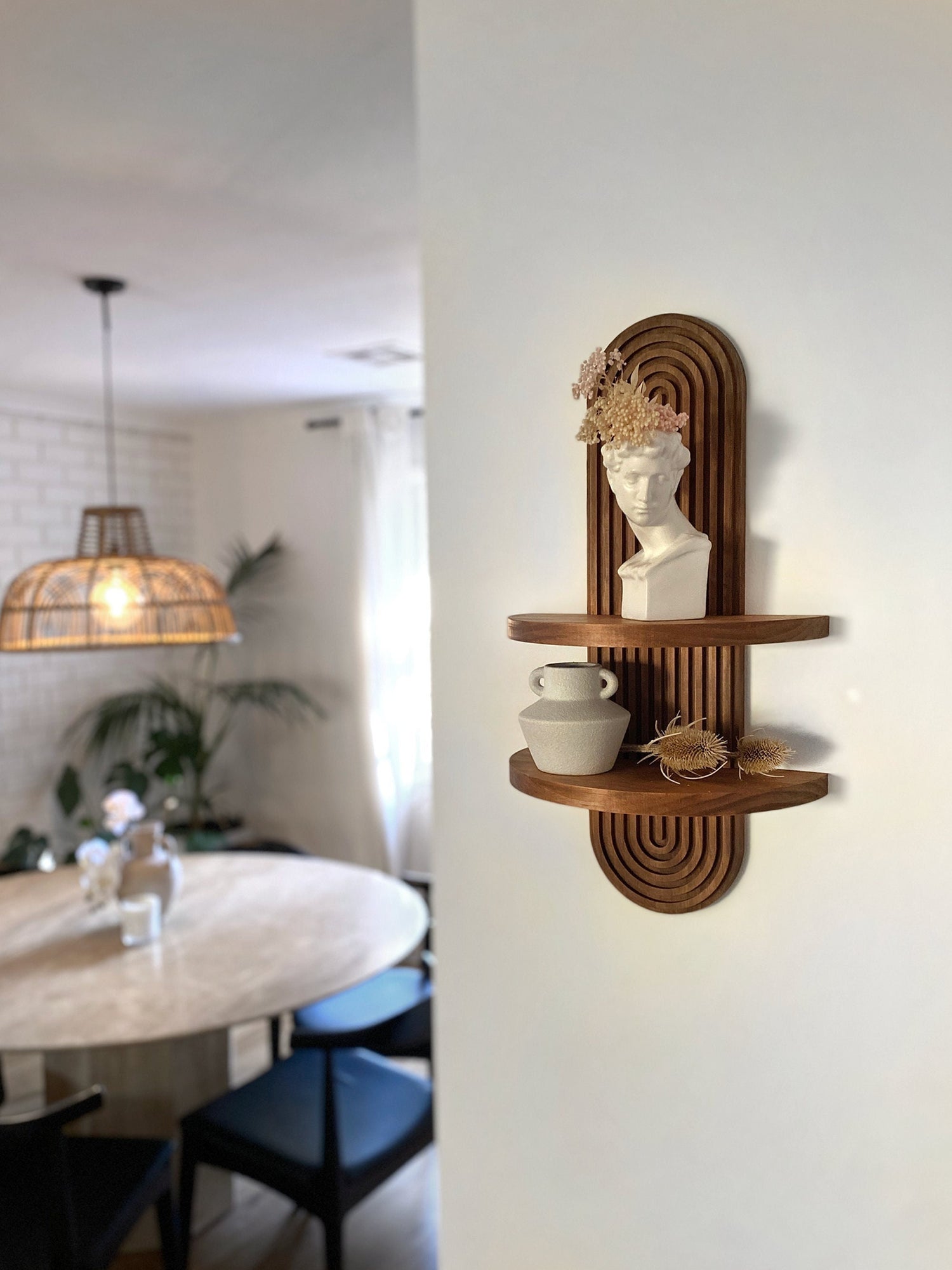 Wooden wall mounted shelf with an arched design, featuring two tiers. The top shelf holds a white bust sculpture with dried flowers, while the bottom shelf holds a ceramic vase and decorative items. Displayed in a modern living space.