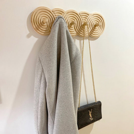 Wooden coat rack with a swirling, spiral design holding a grey coat and a black handbag with a gold chain. The coat rack is mounted on a white wall.