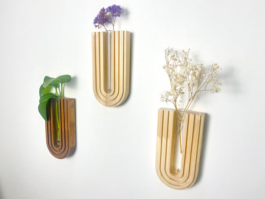 Three mid-century modern wooden wall planters on a white wall. U-shaped design holding a green plant, dried white flowers, and purple flowers. Smooth, natural wood finish, adding a minimalist touch to home decor.
