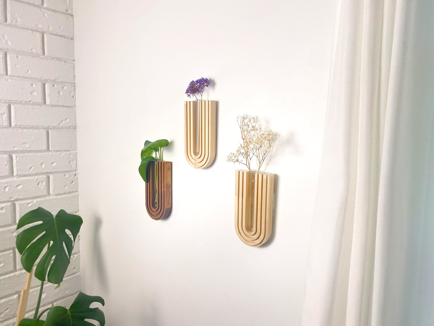 Three mid-century modern wooden wall planters with a U-shaped design on a white wall, holding a green plant, dried white flowers, and purple flowers. The planters have a natural wood finish and are positioned next to a white curtain and a brick wall