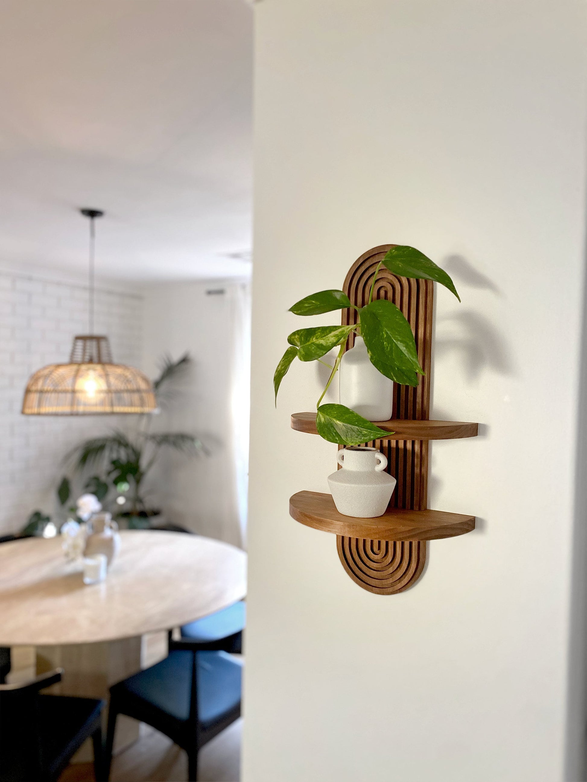 Wooden wall-mounted shelf with an arched design featuring two tiers. The top shelf holds a small potted plant, and the bottom shelf holds a ceramic vase. Displayed in a modern living space with a dining area and wicker pendant light.