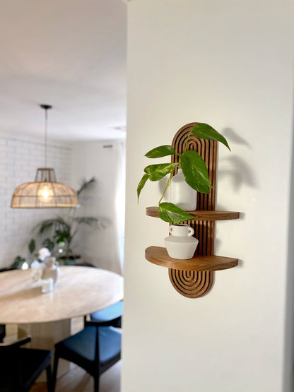 Wooden wall-mounted shelf with an arched design featuring two tiers. The top shelf holds a small potted plant, and the bottom shelf holds a ceramic vase. Displayed in a modern living space with a dining area and wicker pendant light.