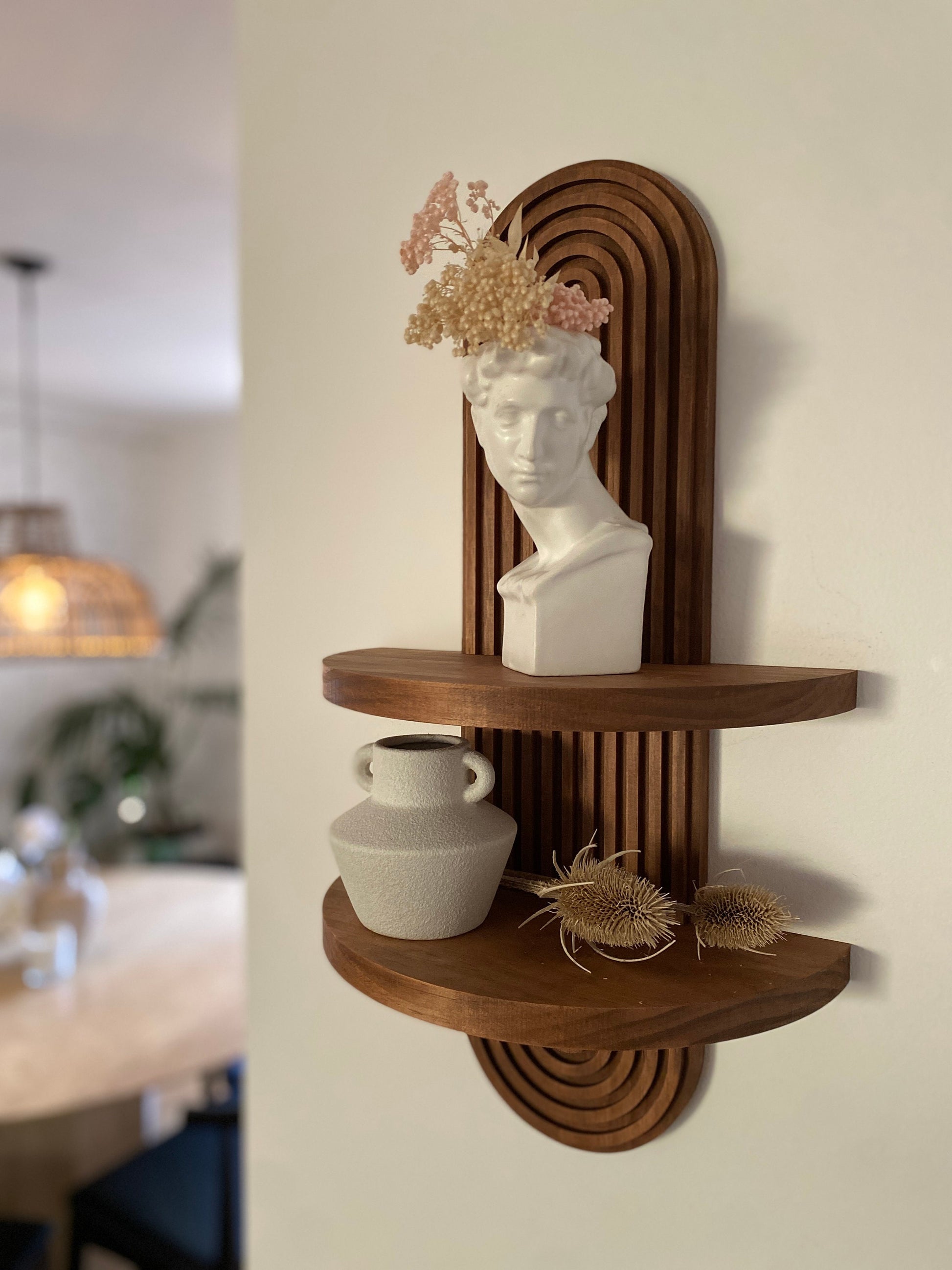 Wooden wall-mounted shelf with an arched design featuring two tiers. The top shelf holds a white bust sculpture with dried flowers, and the bottom shelf holds a ceramic vase and decorative items. Displayed in a modern living space.